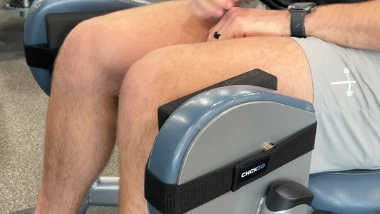 Close-up of a lifter's knees on a seated hip abduction machine, with black CHCK3D Cushie Pads strapped to the machine leg pads with Anchor Bands